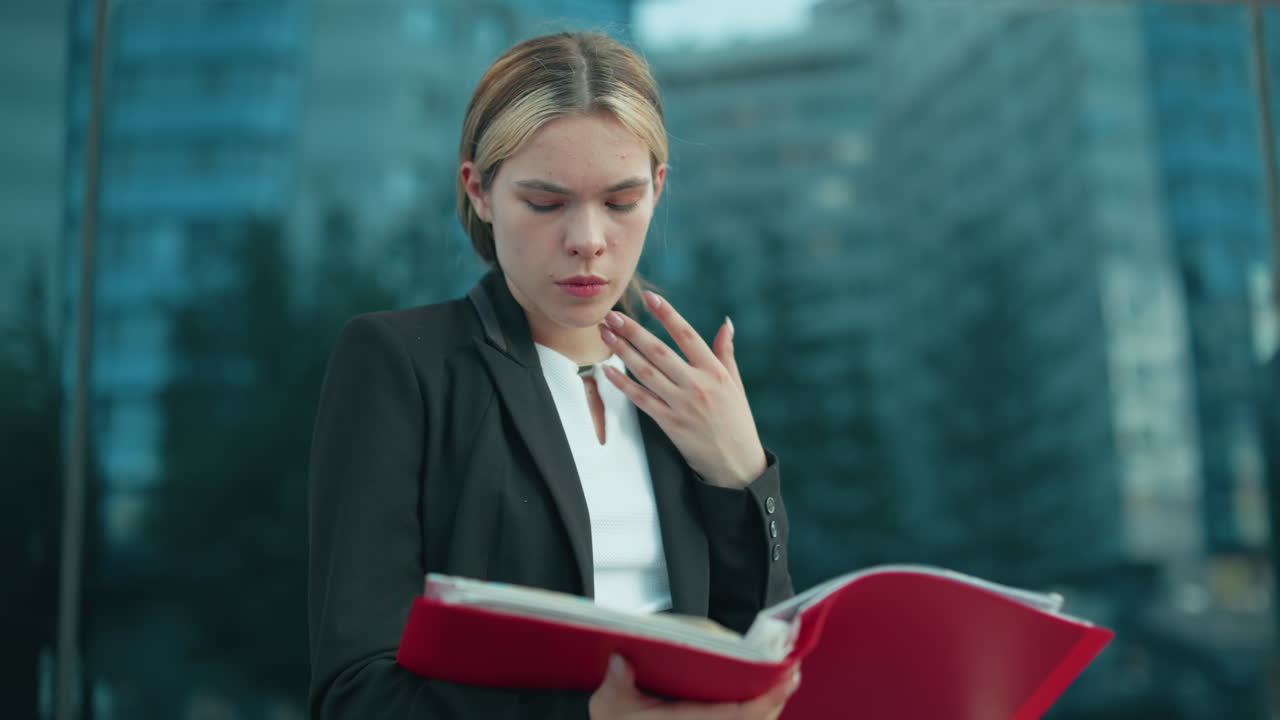 Young professional in black blazer reviewing file attentively near glass residential building reflecting surrounding environment and urban greenery