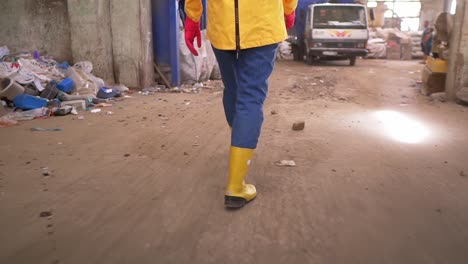 Tracking-rare-view-footage-of-a-girl-in-protective-working-clothes---walking-by-waste-recycling-factory,-huge-area-with-used-bottles,-different-waste-and-white-truck-on-background.-Close-up