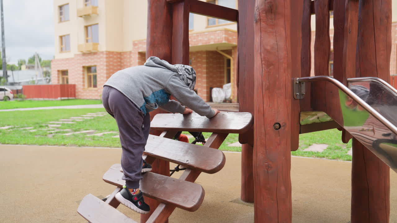 Kid in grey hoodie climbs wooden playground steps, pauses, then steps down carefully while second child in yellow hat walks past, brick building in background