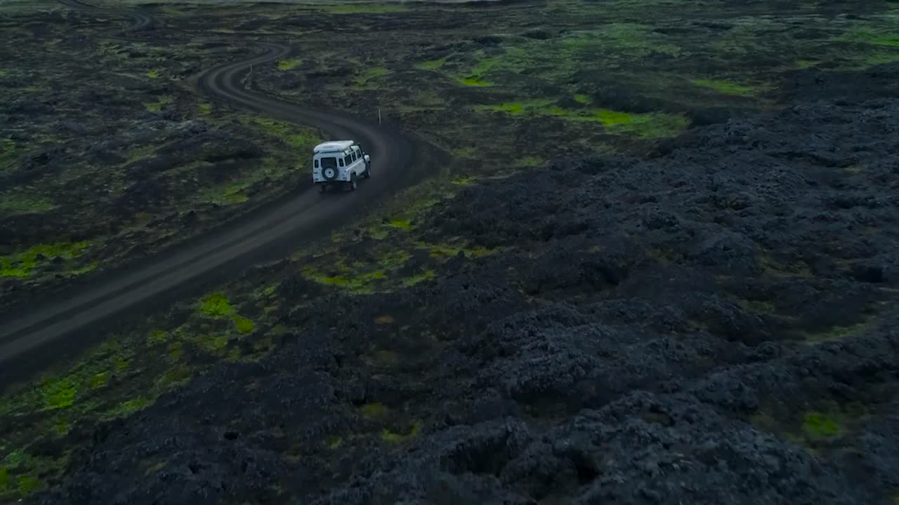 Aerial drone footage of a white 4x4 jeep or a truck driving through and on muddy roads with turns in Iceland or Greenland during a wet and damp evening or dusk weather in the mountains with snow, ice.