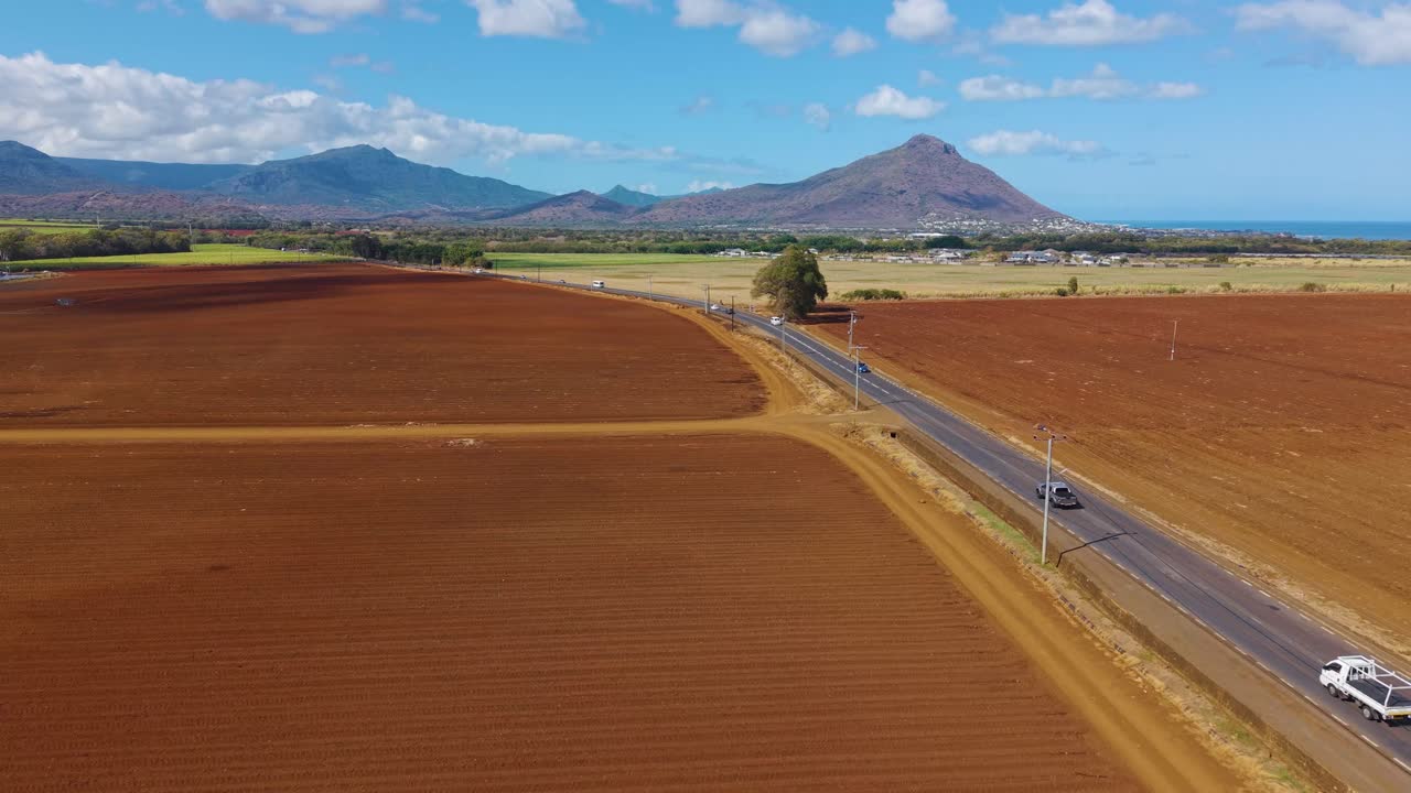 Drone shot of a road on Mauritius island landscape with coastal lagoons, towns, and distant mountains under dramatic tropical clouds and sunlight