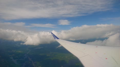 plane window view flying over a hilly forest with clouds below, and beautiful blue skies above