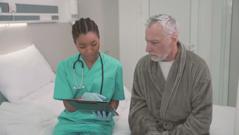 A-Beautiful-Young-American-Female-Doctor-Sitting-On-The-Edge-Of-A-Hospital-Bed-With-An-Old-Patient