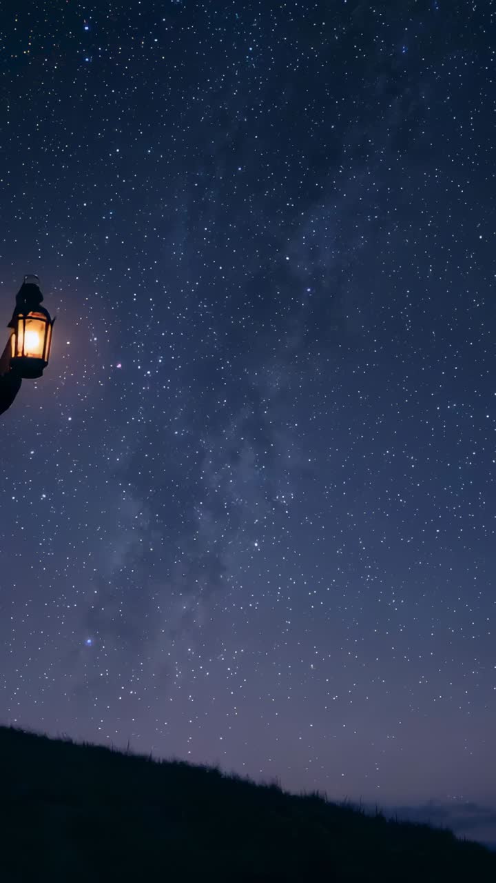 Vertical video: Shifting forward woman in coat raising lantern on ridge under Milky Way, copy space