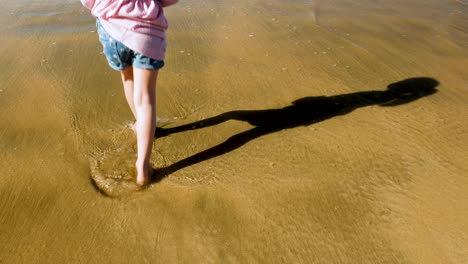 Schatten-Eines-Jungen-Mädchens,-Das-Winkt,-Während-Es-Im-Knöcheltiefen-Wasser-Am-Strand-Steht