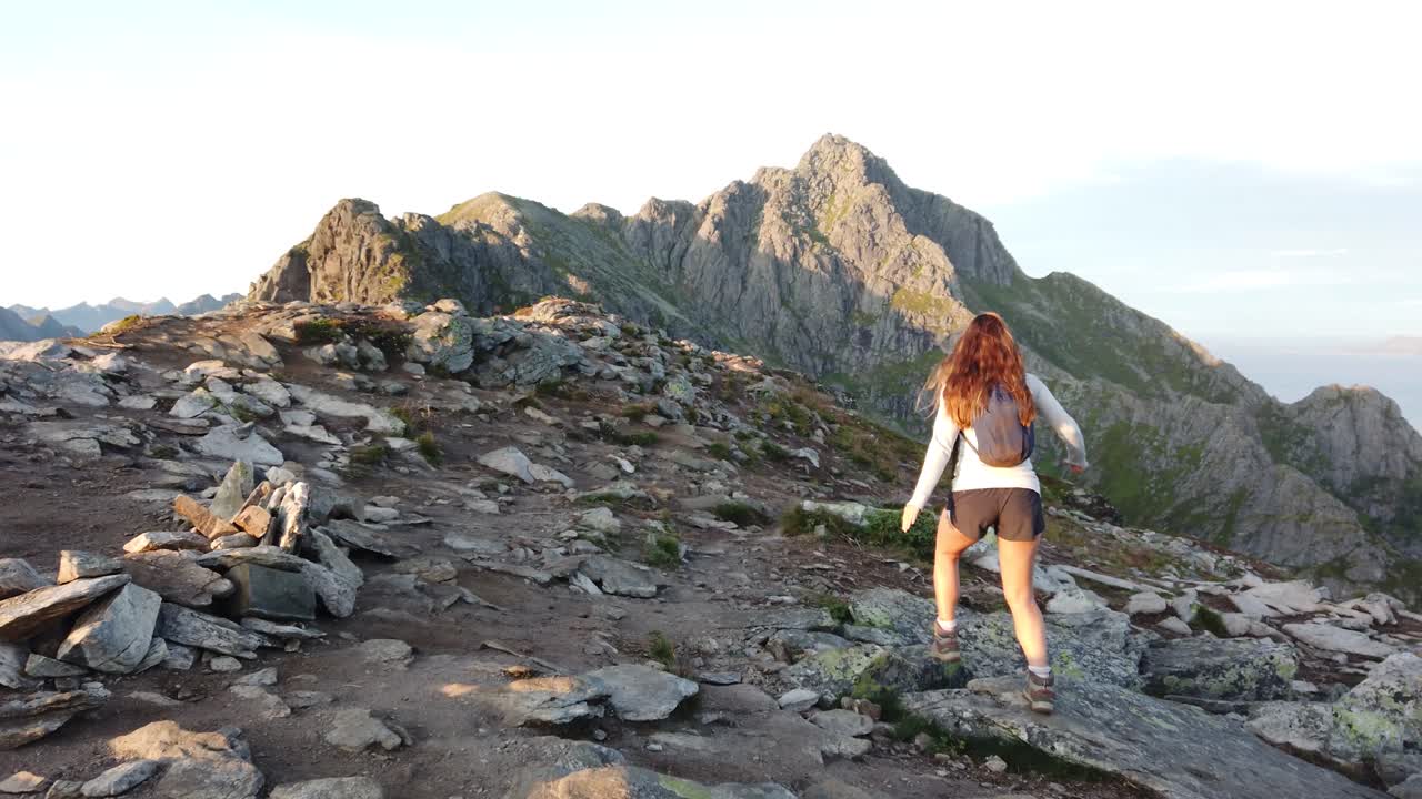 una mujer joven con cabello largo y castaño camina sola sobre rocas y piedras en la cima de la montaña festvågtind en lofoten durante el sol de medianoche