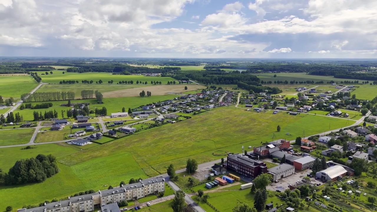 Luftaufnahme mit Drohne über die Landschaft von Sigulda in Lettland, die grüne Felder, Wohngebiete, Landstraßen, kleine Bauernhöfe und einen fernen Horizont unter bewölktem Sommerhimmel zeigt