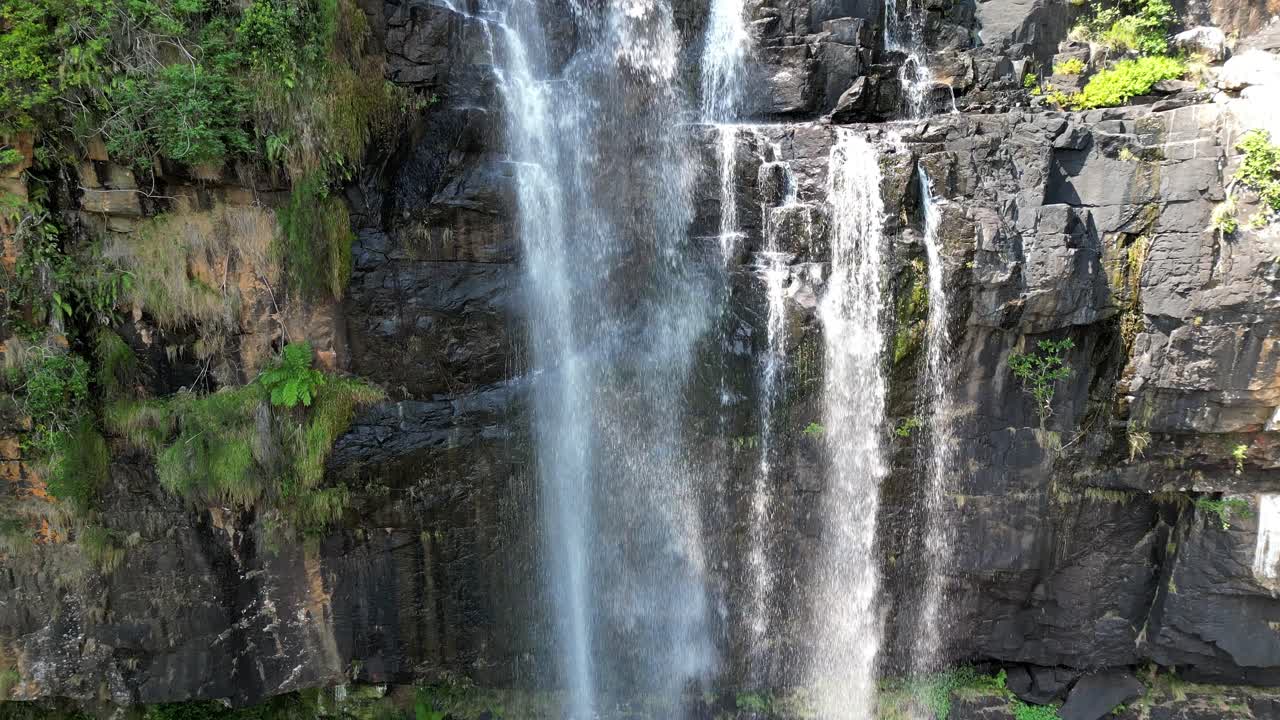 Tall alpine waterfall drops in thin streams along dark rock wall before landing in clear emerald pool surrounded by moss and steep cliff creating bright peaceful mountain scene in Bavaria Germany
