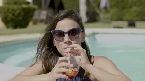 vista frontal de una mujer feliz con traje de baño azul y gafas de sol, bebiendo un cóctel en la piscina