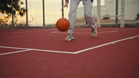 Close-up-shot-of-a-girl-in-white-pants-hitting-an-orange-basketball-on-a-red-basketball-court-early-in-the-morning.-A-girl-in-white-pants-does-exercises-and-practices-handling-an-orange-basketball-in-the-summer