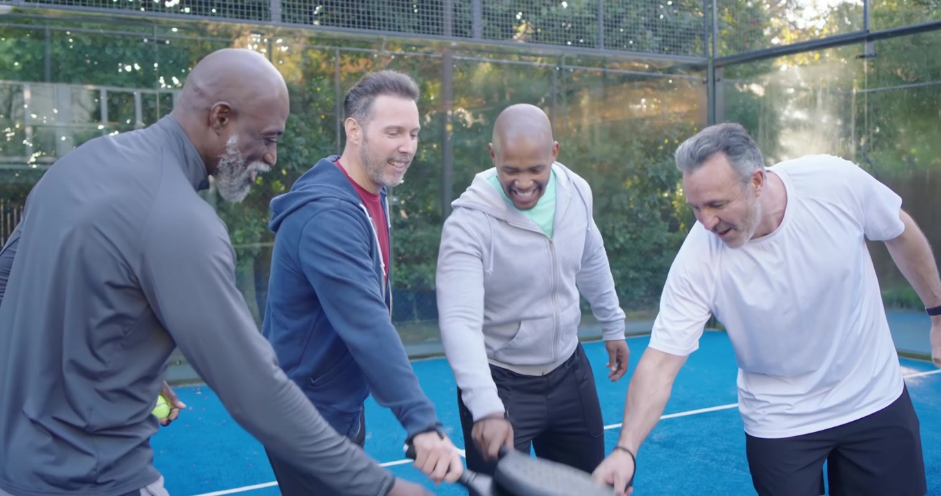 Diverse male friends chatting, passing yellow balls with paddles on blue court huddling before play