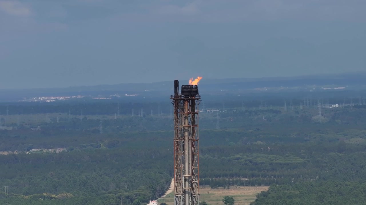 Altas llamas danzan desde una imponente torre de combustión en una refinería de petróleo rodeada de exuberantes paisajes