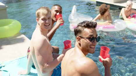 portrait of happy diverse friends playing and drinking drinks at pool party in summer