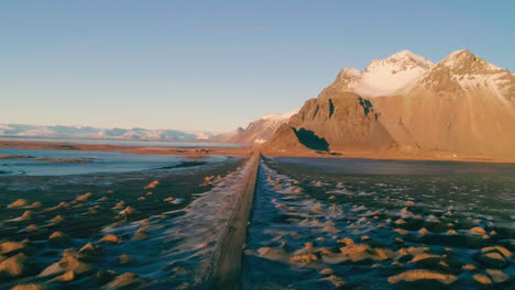 Long-road-leading-to-Vestrahorn-golden-sunrise-Mountain-across-low-tide-Stokksnes-beach,-Aerial-view