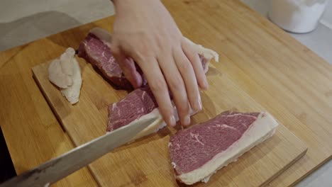 chef cuts off fat from the steak with steel kitchen knife on a wooden cut board