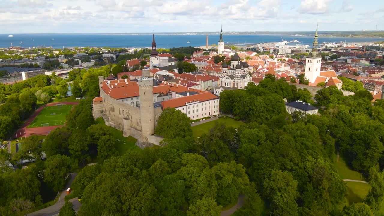 Aerial drone footage orbiting and revealing Tallinn Old Town medieval city walls, parliament building and Toompea Hermann tower with a Estonian flag on it. Tallinn harbor visible in the back with sea