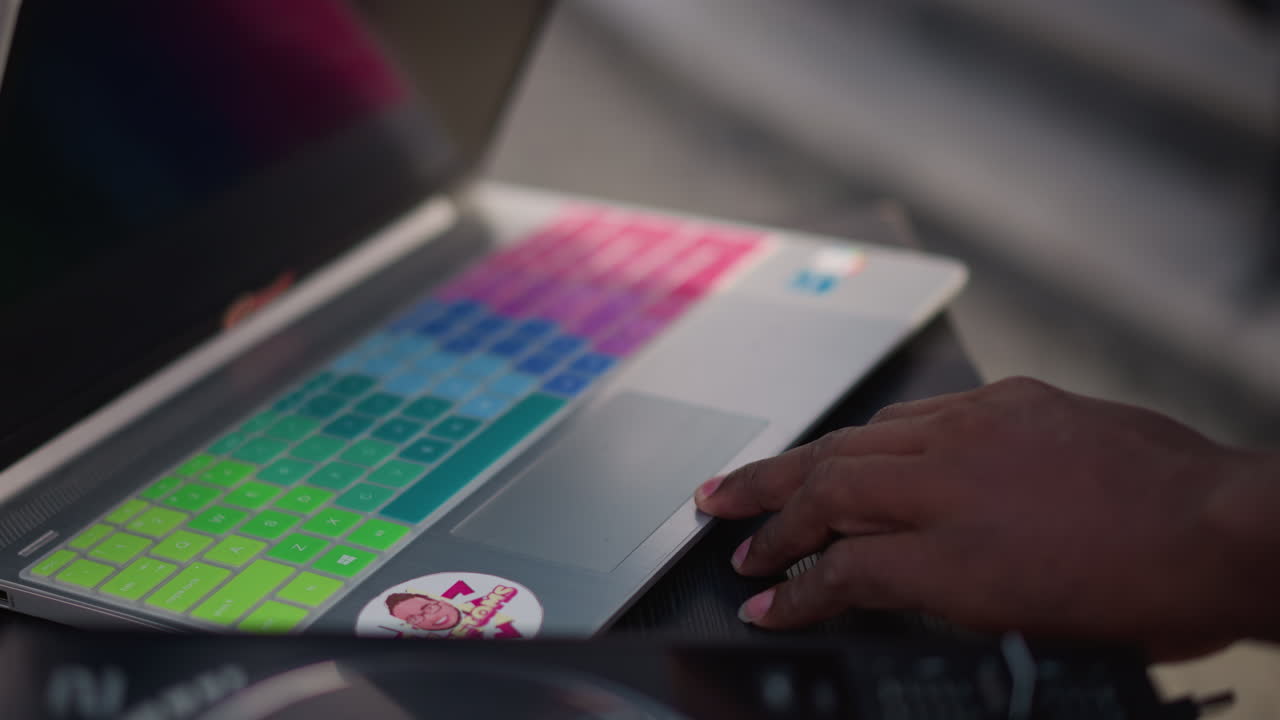 DJ di colore che prepara la cover colorata del laptop, mani che girano la decalcomania per rivelare tastiera retroilluminata con pad al neon, allestimento da tavolo di strada all'aperto, artista che regola i controlli della traccia, energia concentrata e movimento ritmico