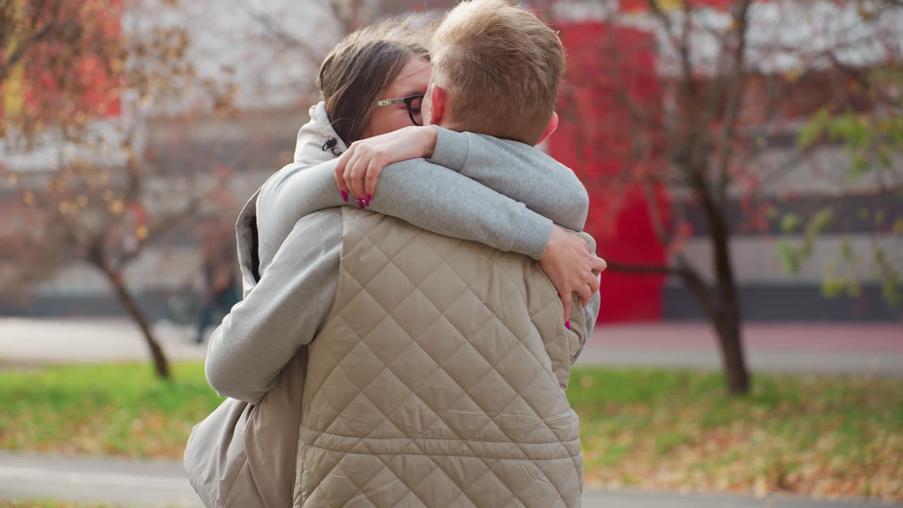 Lovers embrace warmly while sharing kiss in matching outfits under autumn trees with gentle breeze swaying leaves as people walk in distance