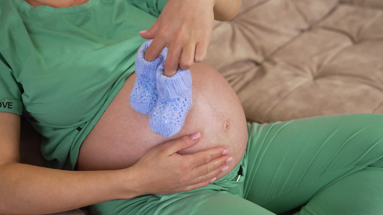 Young future mother holding knitted shoes. Pregnant woman with a big bare belly playing with little blue socks at home. Pregnancy.