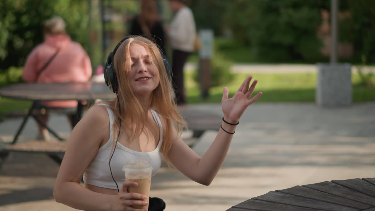 Eine junge Frau hört im Freien mit einem Eiskaffee Musik. Sie sitzt an einem Tisch im Park, trägt große Kopfhörer und lächelt lässig, während sie im sommerlichen Sonnenlicht zur Melodie schwingt. Bäume und Picknickbänke im Hintergrund.