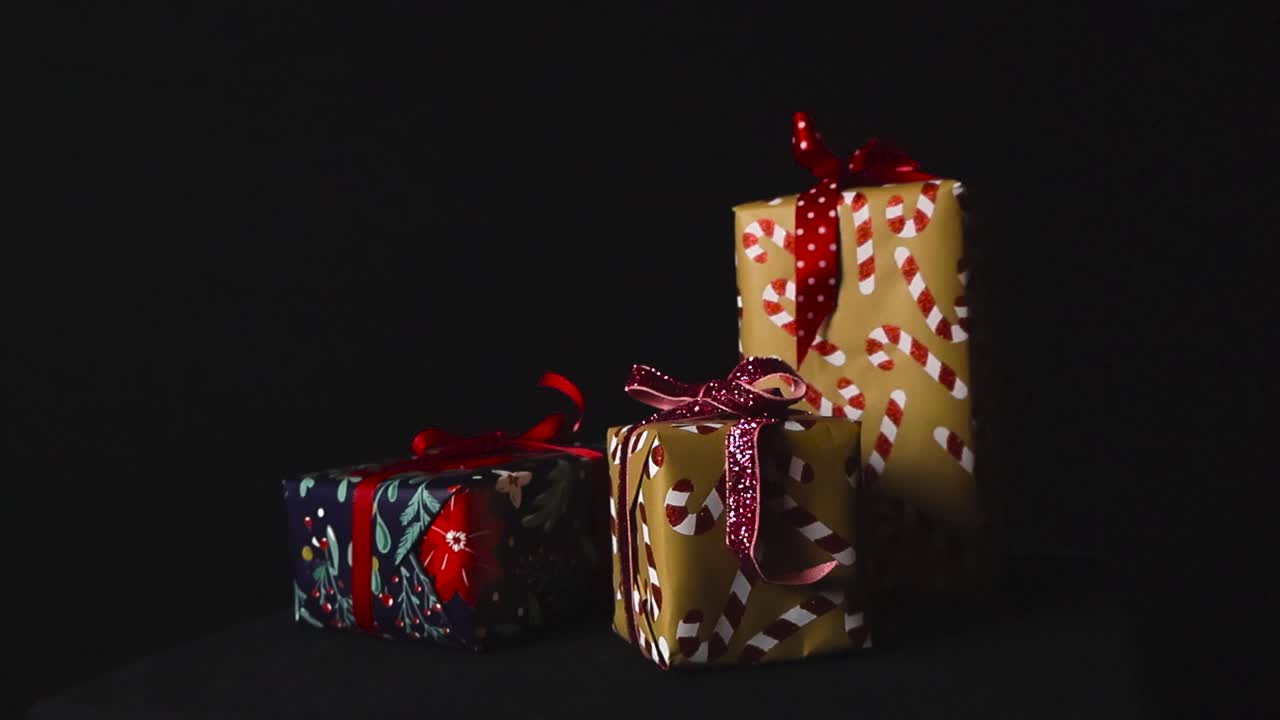 Closeup gliding around beautifully wrapped Christmas presents displayed on dark surface against black backdrop in studio setting under soft light casting shadows. Rosettes fall on gifts in slow motion