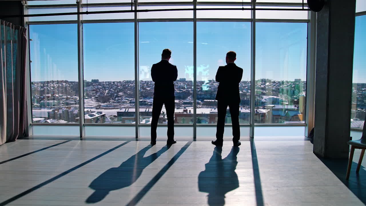 Full-length portrait of business people. Male entrepreneurs in elegant suits standing in modern office and talk about business while looking at the view. Panoramic windows view on city