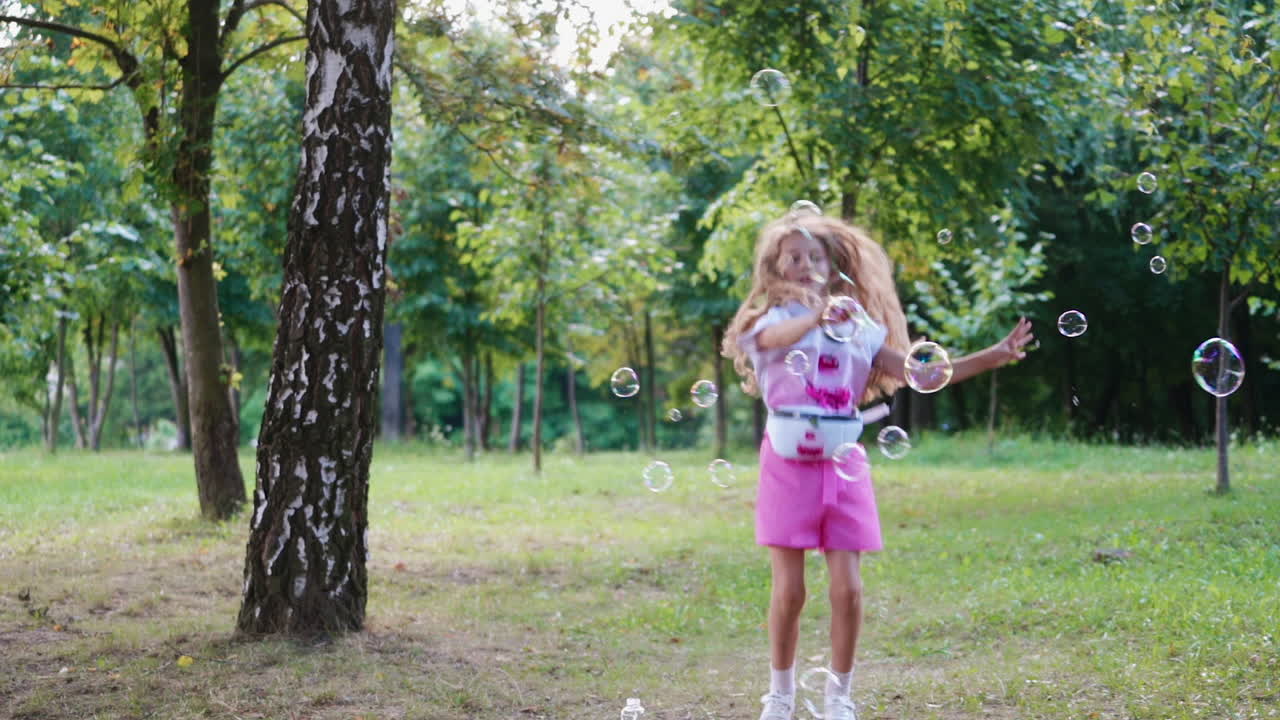 Happy girl playing with bubble blower in the park. Beautiful child jumping joyfully waving wand with soap bubbles on the natural background.