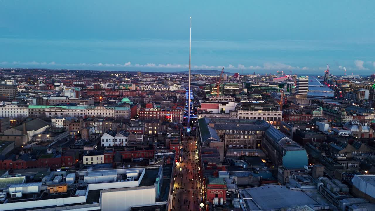 Luftfoto av landskap tatt med drone i bred vinkel, flygende mot spirlandemerket i Dublin sentrum i skumringen under den blå timen, vinterlig solnedgang med irsk bybilde og opplyste bygninger og trafikkerte veier nedenfor