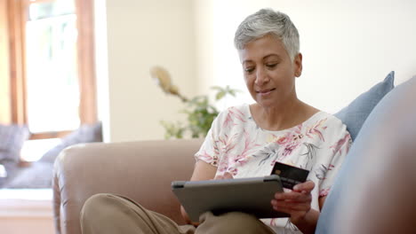 mujer biracial senior feliz en el sofá con tarjeta de crédito y usando tableta en casa, cámara lenta
