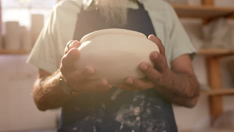 mid section of biracial potter with long beard holding bowl in pottery studio, slow motion