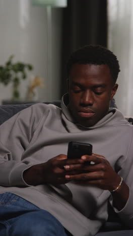 vertical video of young man sitting on sofa at home messaging or browsing on mobile phone