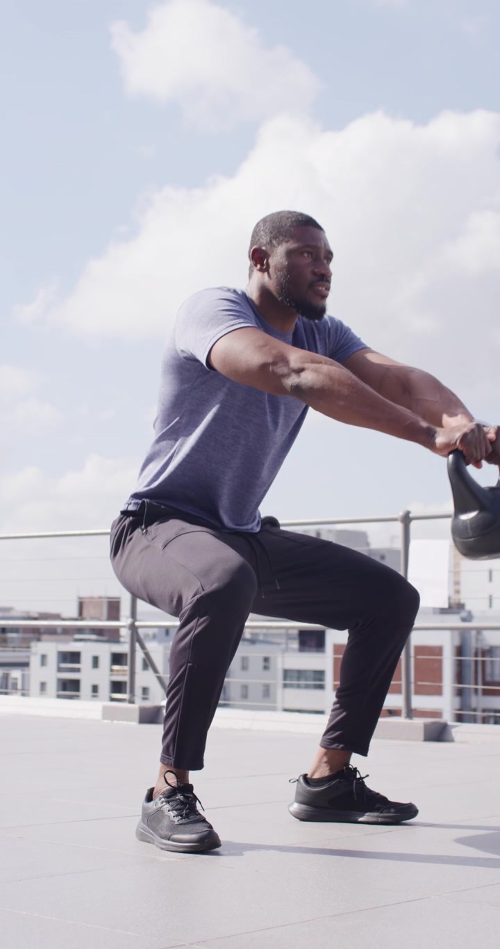 Video verticale: Uomo afroamericano di mezza età che si muove, si accovaccia ed estende un kettlebell su un tetto.