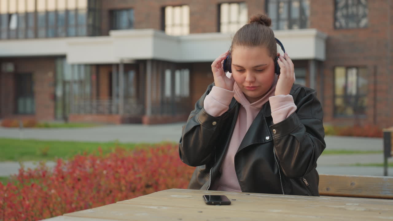 Young woman seated outdoors puts headset on while reaching for phone on wooden table with residential building and red shrubs in background during calm windy day in urban park setting