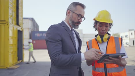 ejecutivo de mediana edad, gerente o inversor y ingeniera femenina profesional del proyecto hablan detalles o proceso de carga y envío de carga en la tableta en el patio de carga de contenedores. concepto de cooperación en negocios. panorámica alrededor de la toma