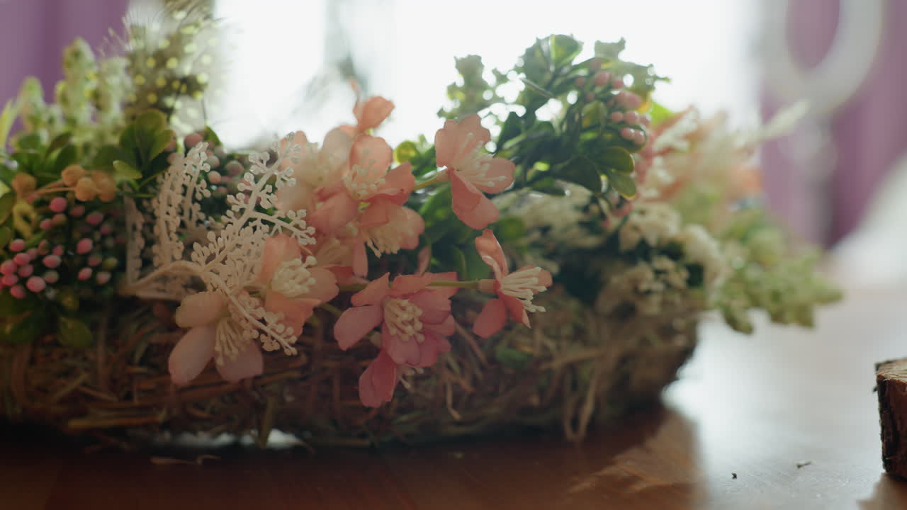 Hender som jobber med rustikk blomsterkrans dekorert med rosa blomster, grønne blader og naturlige kvister på et trebord. En kreativ håndverker arrangerer håndlaget dekorasjon med mose og sesongplanter