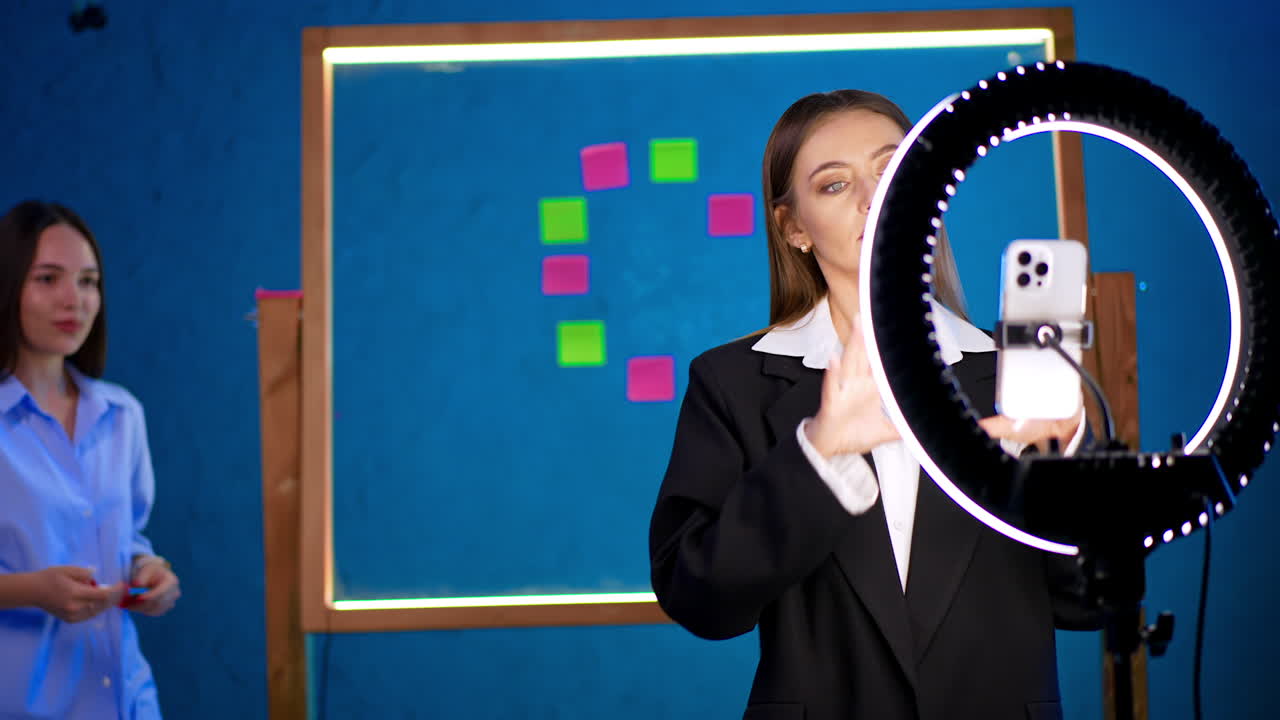 Woman in black jacket stands at tripod with camera and ring light. Lady talks and waves her hands actively. A girl comes up and talks to camera too. Blogging concept.