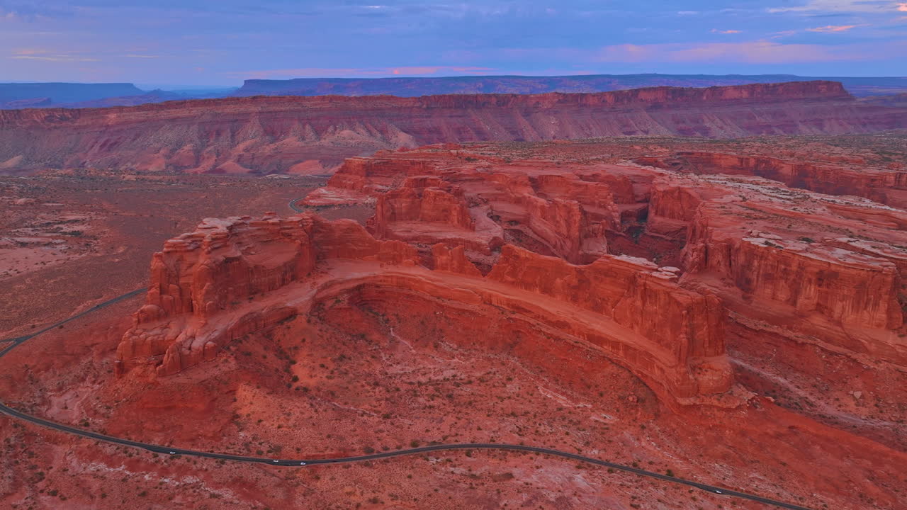 Distancing from the spectacular red rocks with flat tops. A highway crosses the desert. View on the Arches National Park, Utah, USA