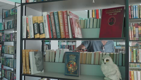 young man in casual clothing standing near tall bookshelves in library, partially visible while browsing and looking around at rows of colorful books in quiet indoor environment
