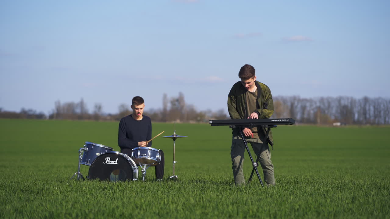 Musicians performing music playing the drums and piano. Members of boys' band play music and dance in the field. Highway with cars at the backdrop.