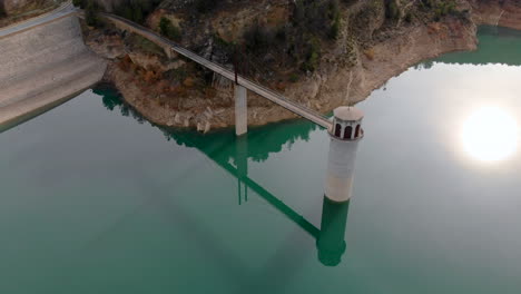 Un-Dron-Orbita-Alrededor-De-Una-Torre-Unida-A-Una-Presa-Sumergida-En-Agua-Turquesa-Del-Embalse