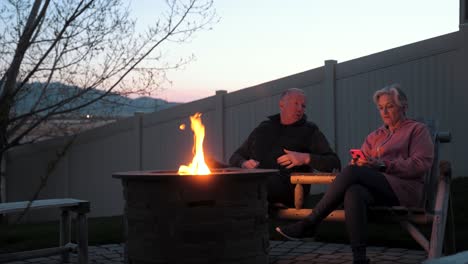 senior husband asks his wife to put her smartphone down and pay attention as they sit at a backyard fire pit