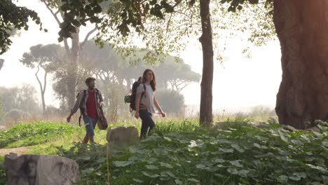 young happy couple backpackers tourists walking in the nature along dirt road in aqueduct park ruins in rome countryside on misty morning with guitar and sleeping bag slow motion