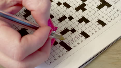 closeup of a woman with pink fingernails working on a crossword puzzle with a black pen with one answer filled in so far