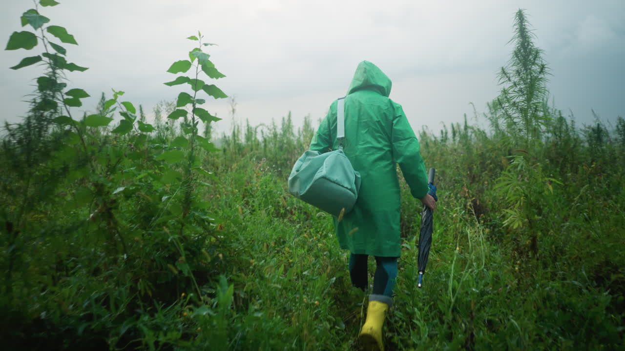 Back view of woman in green raincoat carrying mint-colored bag and umbrella, wearing yellow rain boots, walking cautiously along a path surrounded by tall greenery and plants