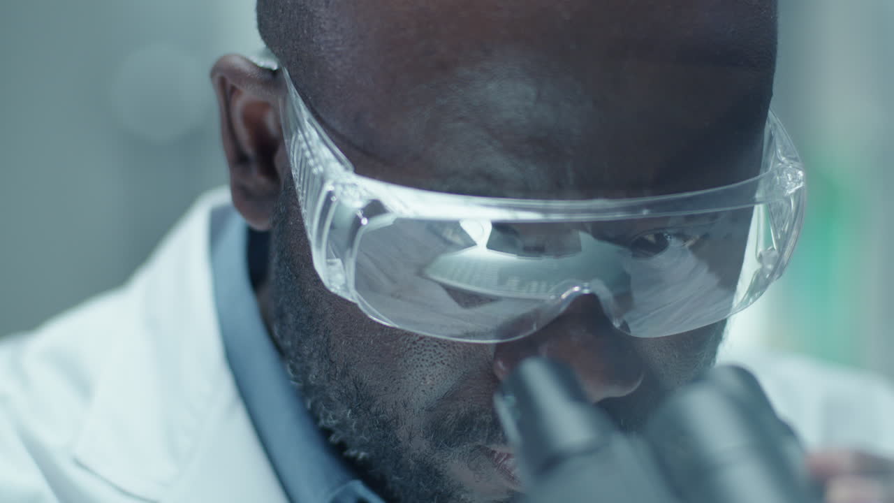 Black Chemist in Safety Glasses Looking through Microscope in Laboratory