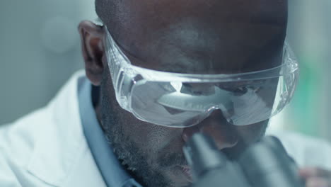 black chemist in safety glasses looking through microscope in laboratory