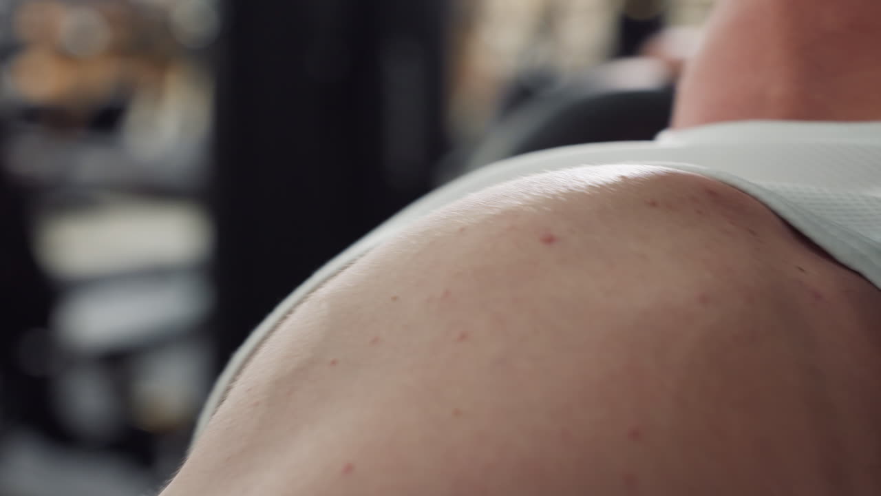 Close up of muscular young man shoulder and upper arm straining under white singlet on chest press machine muscles flexing sweat sheen highlighting effort and form during intense gym workout