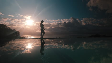 woman walking beside calm infinity pool with her silhouetted reflection against the clouds and sun