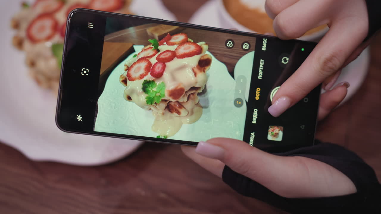 Close-up view of hand holding smartphone to capture delicious waffle dessert topped with sliced strawberries and cream, placed on white plate and saucer on a brown wooden table