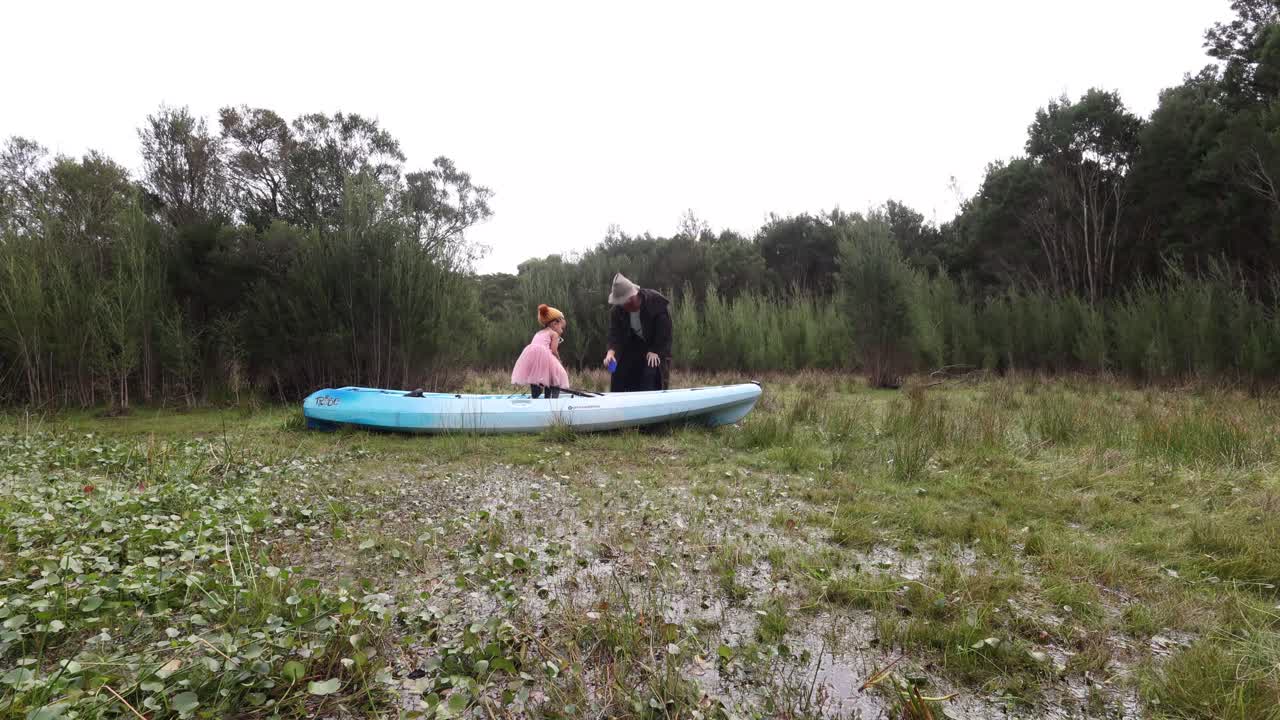 A man in a trenchcoat drinks tea out in a swamp with his daughter in a kayak.